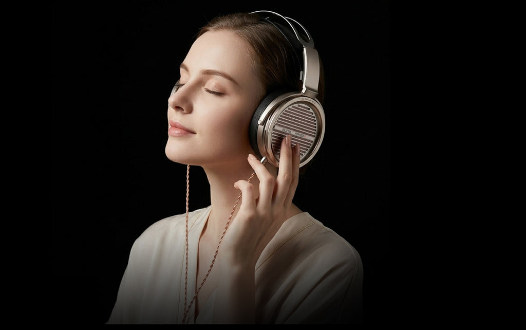 A woman with her eyes closed wears over-ear headphones and gently touches the earcup, set against a solid black background.