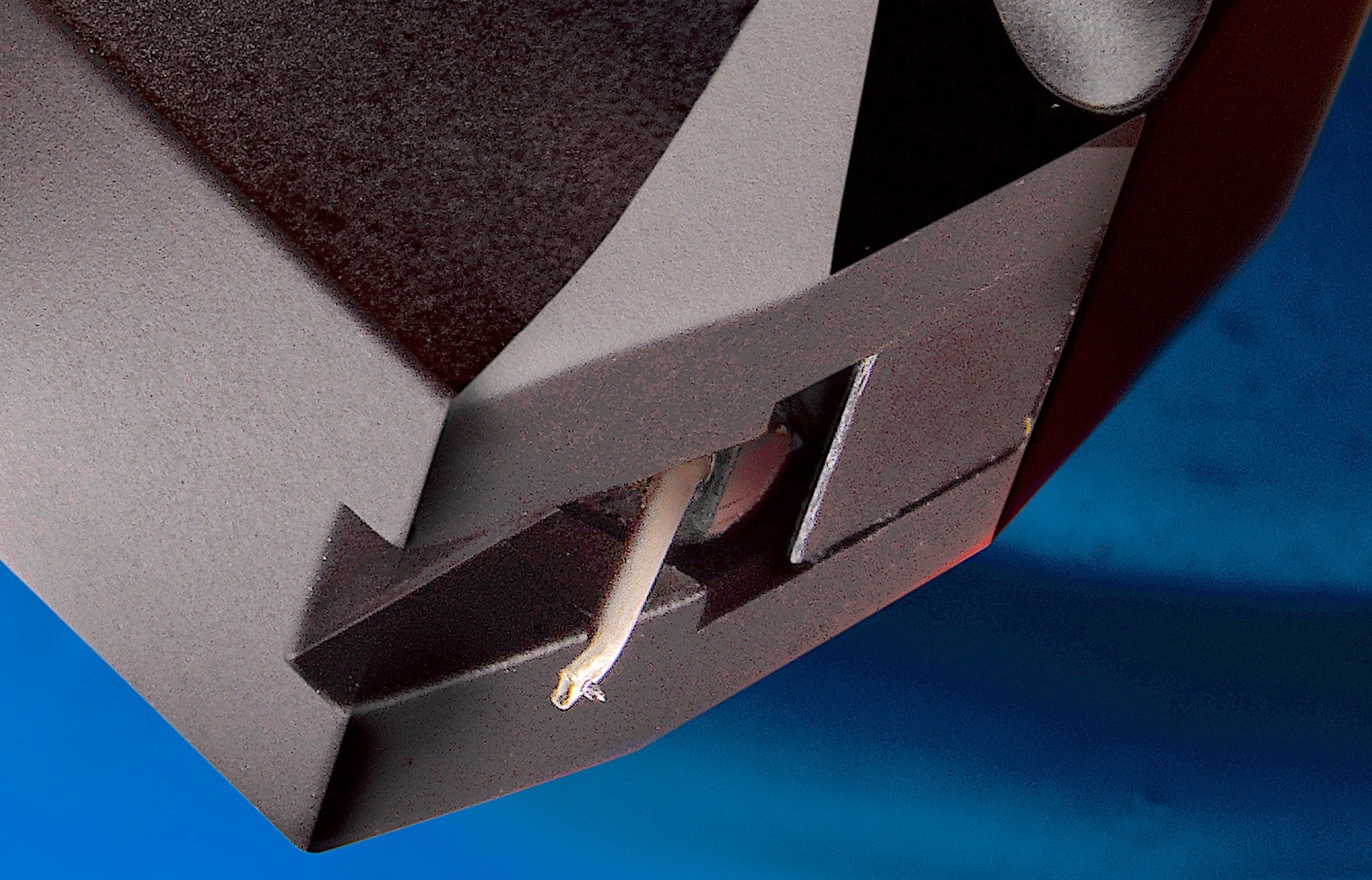 Close-up detail of a dark gray or black audio component or speaker housing with a visible white wire, set against a bright blue background.