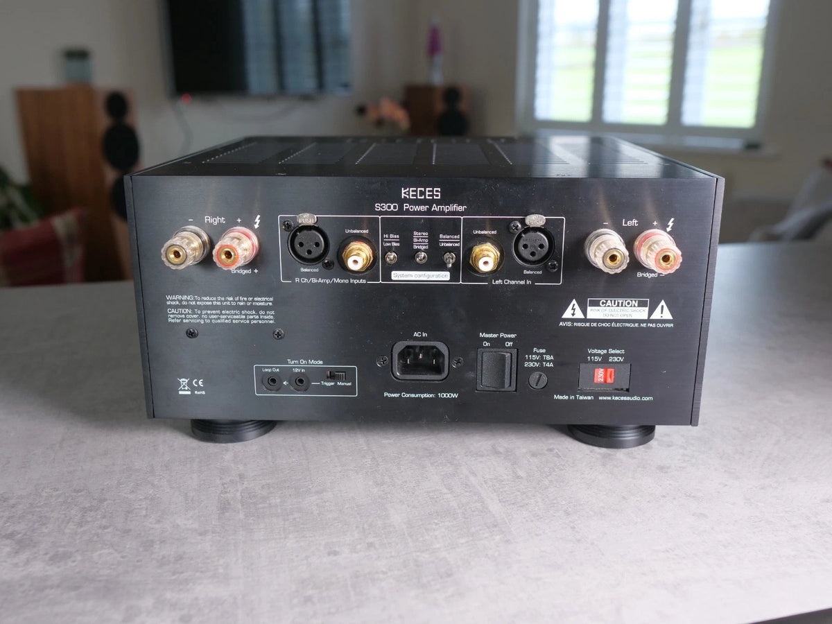 A black KECES S300 power amplifier receiver sitting on a gray tabletop in a home environment, showing its rear panel with various audio input and output connectors.