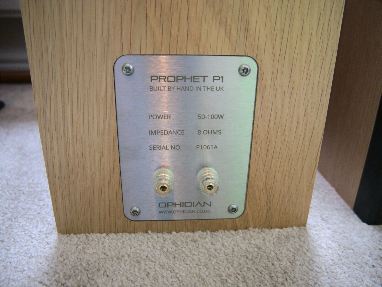 Close-up of the rear terminal plate of an Ophidian Prophet P1 speaker, showing two RCA connectors on a brushed silver metal plate mounted on a light wood cabinet, set on a beige carpet.