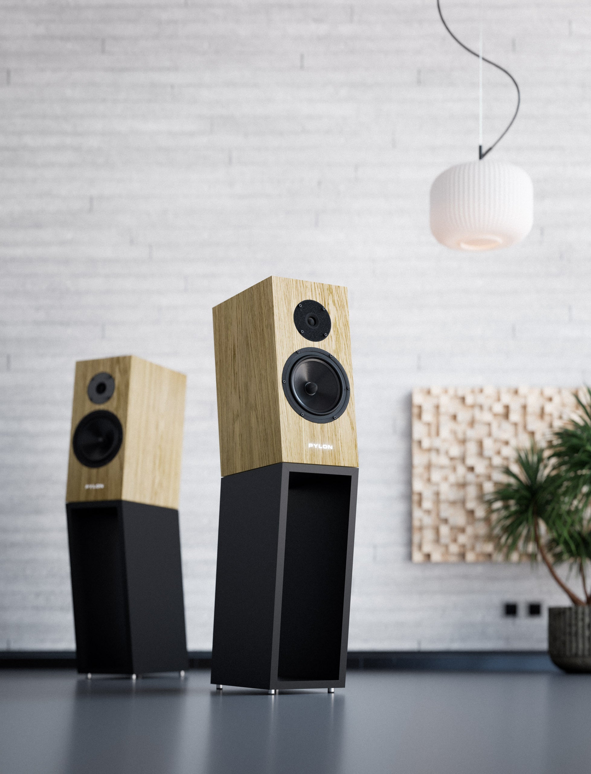 Two modern floor-standing speakers with light wood grain tops and black lower cabinets, displayed in a minimalist room setting with a white brick wall, pendant light, and potted plant.
