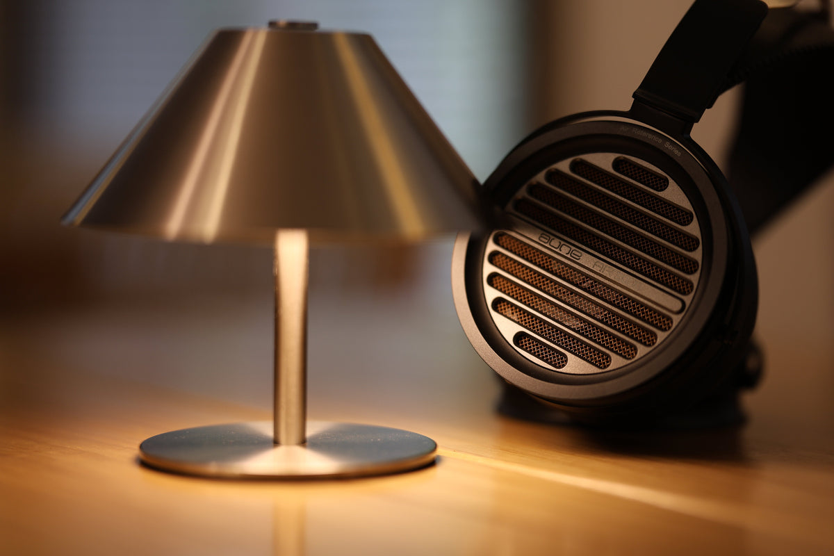 A modern table lamp with a brushed metal shade and base sits on a wooden surface, casting a warm glow next to a pair of aune ar3000 over-ear headphones.