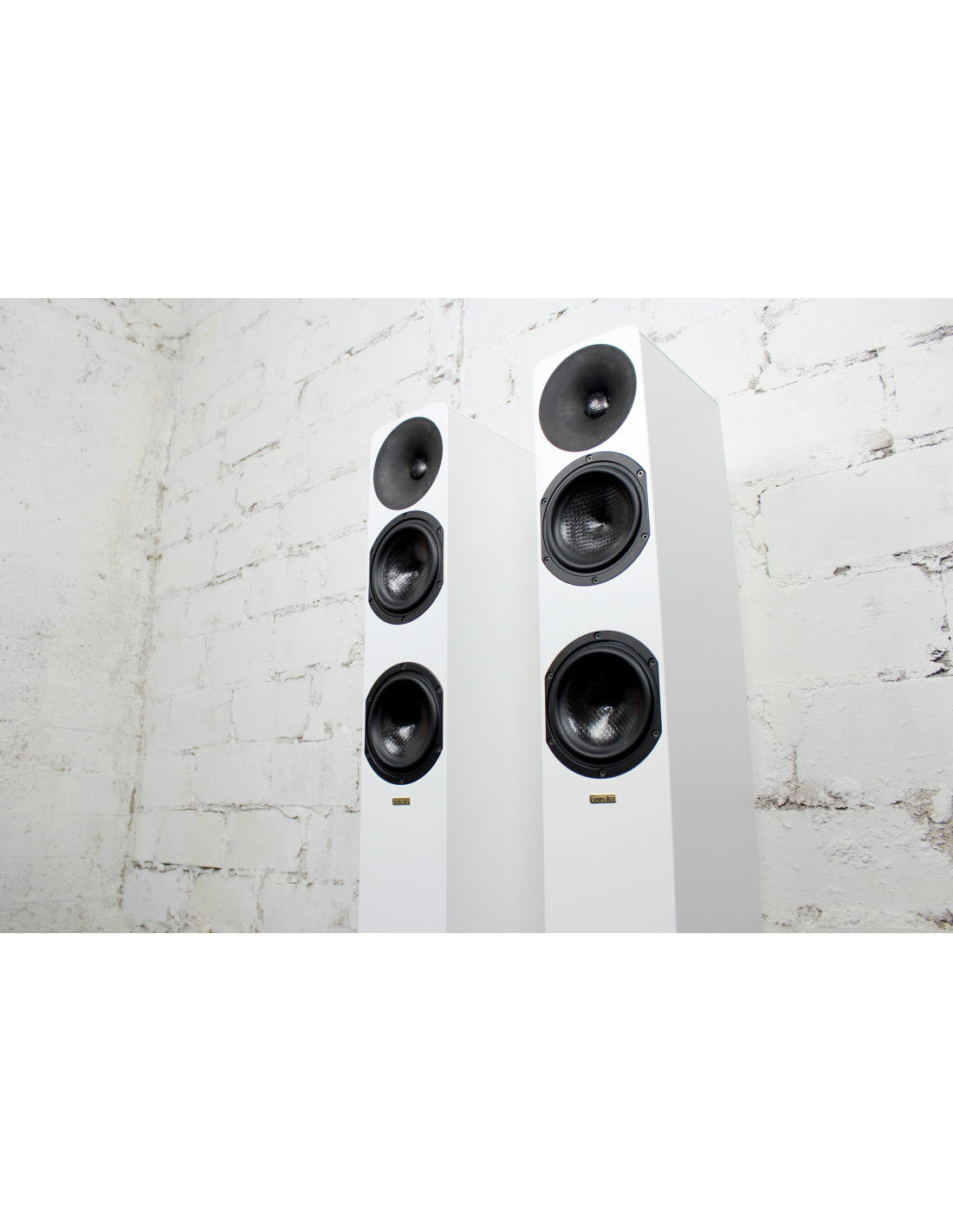 Two white Aurelia Ambera floorstanding speakers with black drivers, set against a white brick wall background.