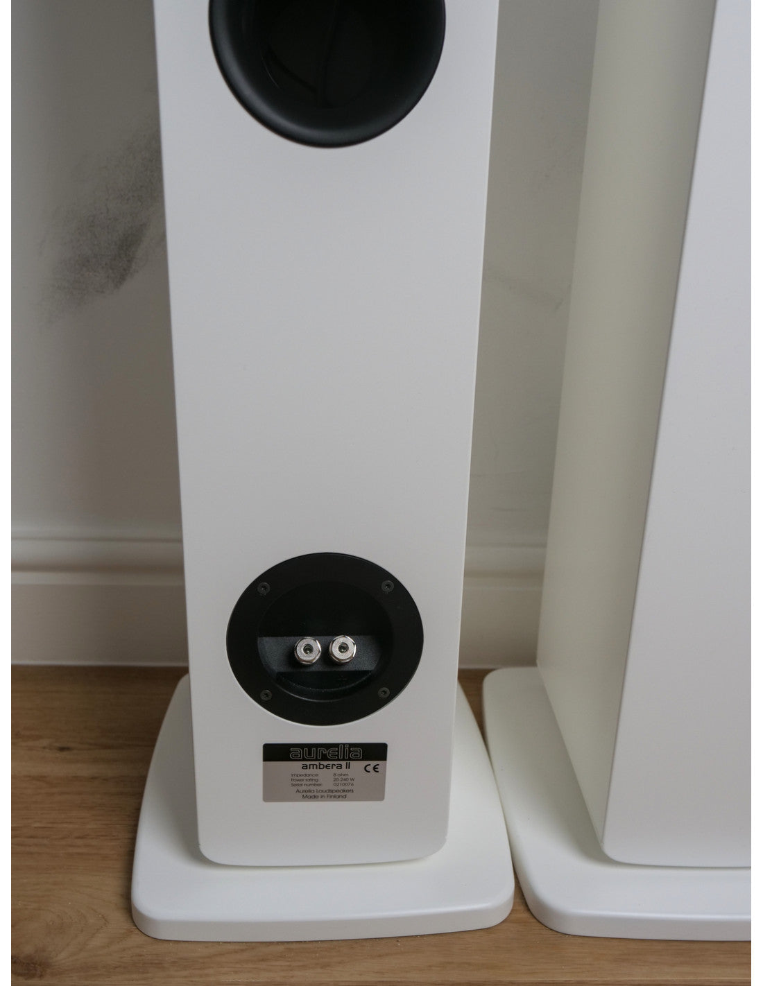A close-up of a white Aurelia Ambera II Floorstanding Loudspeakers  standing on a wooden floor against a white wall, showing the rear port and terminal details.