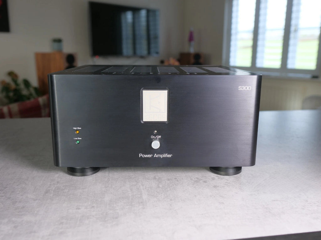 A black power amplifier speaker system with a brushed metal front panel, standing on a gray tabletop in a blurred living room setting.