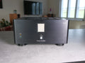 A black power amplifier speaker system with a brushed metal front panel, standing on a gray tabletop in a blurred living room setting.